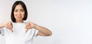 Asyalı bir kadının başparmaklarını göstermesi, smth 'den hoşlanmaması, hayal kırıklığına uğramış görünmesi, beyaz arka planda durması.