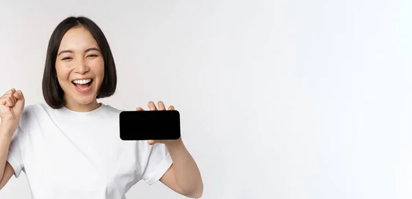 Enthusiastic asian girl scream in joy, showing horizontal smartphone screen, mobile phone display, standing over white background — Stock Photo, Image