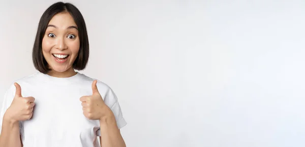 Glückliche junge Koreanerin zeigt Daumen nach oben und lächelt, gibt positives Feedback, empfiehlt etw., steht über weißem Hintergrund — Stockfoto