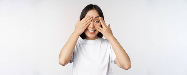 Portrait of asian woman covering eyes, waiting for surprise blindfolded, smiling and peeking at camera, standing over white background