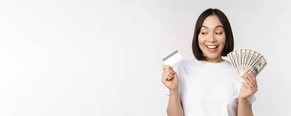 Porträtt av asiatisk kvinna leende, innehav av kreditkort och pengar kontanter, dollar, stående i t-shirt över vit bakgrund — Stockfoto