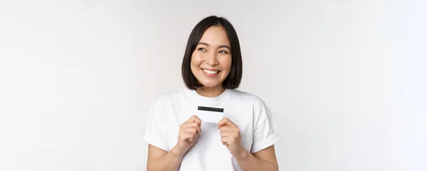 Lächelnde Koreanerin zeigt Kreditkarte mit glücklichem Gesicht und steht im T-Shirt vor weißem Hintergrund — Stockfoto