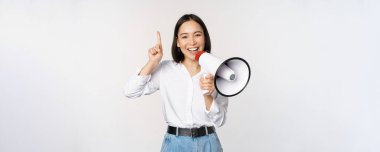 Gülümseyen mutlu Asyalı kız megafonla konuşuyor ve işaret ediyor, indirimli tanıtım duyurusu yapıyor, üzerinde reklamını yapıyor, beyaz arka planda duruyor.