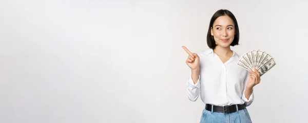笑顔の若い現代アジアの女性は、バナー広告を指して、現金のお金のドルを保持し、白い背景の上に立って — ストック写真