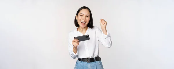 Glad asiatisk kvinna gör vinnardans, triumferande, vinna på mobiltelefon videospel, hålla smartphone horisontellt läge och fira, vit bakgrund — Stockfoto