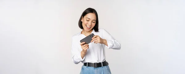 Ung koreansk kvinna, asiatisk flicka spelar mobilt TV-spel på smartphone, tittar på horisontell telefonskärm, står över vit bakgrund — Stockfoto