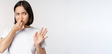 Asyalı kız iğrenmiş görünüyor, kötü kokulu ürünü reddediyor, tiksinti ve korkuyu önlüyor, beyaz arka planda duruyor.