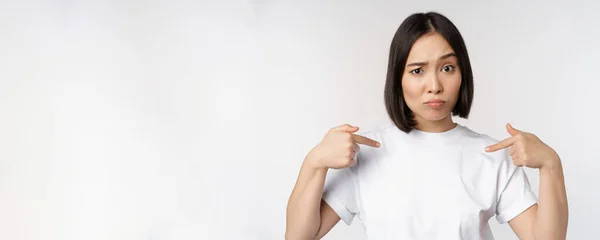 Joven mujer asiática señalándose a sí misma con incredulidad, siendo elegida, sorprendida por su candidatura, de pie sobre fondo blanco — Foto de Stock