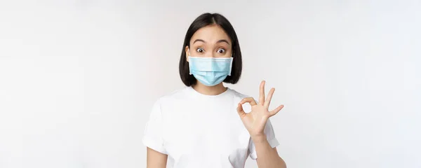 Covid-19, hälso- och sjukvårdskoncept. Imponerad asiatisk kvinna i medicinsk mask, ser förvånad och visar ok, okej tecken, står över vit bakgrund — Stockfoto