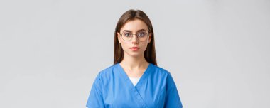 Sağlık çalışanları, ilaç, sigorta ve covid-19 salgını konsepti. Genç bayan stajyer, hemşire ya da doktor, önlüklü ve gözlüklü, ciddi bir ifadeyle kameraya bakıyor, gri arkaplan