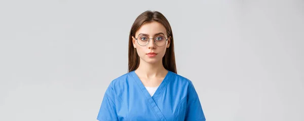 Sjukvårdspersonal, medicin, försäkring och covid-19 pandemi koncept. Ung kvinnlig praktikant, sjuksköterska eller läkare i skrubb och glasögon, tittar på kamera med allvarliga uttryck, grå bakgrund — Stockfoto