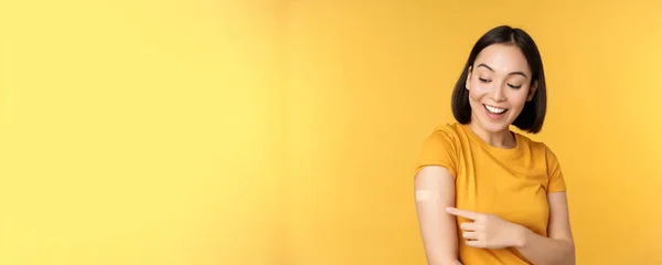 Impfung und Covid-19-Pandemiekonzept. Glückliches und gesundes asiatisches Mädchen zeigt nach Impfung gegen Coronavirus mit Pflaster auf ihre Schulter, gelber Hintergrund — Stockfoto