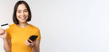 Güzel gülümseyen Asyalı kız kredi kartı ve cep telefonu kullanıyor, akıllı telefondan ödeme yapıyor, beyaz arka planda sarı tişörtle dikiliyor.