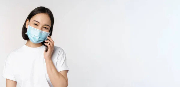 Health and covid-19 concept. Smiling asian girl in medical face mask talking on phone, answer mobile call, standing over white background — Stock Photo, Image