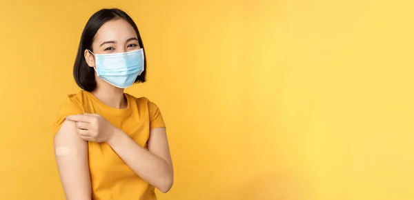 Vaccinatie en covid-19 pandemie concept. Glimlachende Aziatische vrouw met medisch gezichtsmasker, die haar schouder laat zien met pleister na vaccinatie met coronavirus, gele achtergrond — Stockfoto