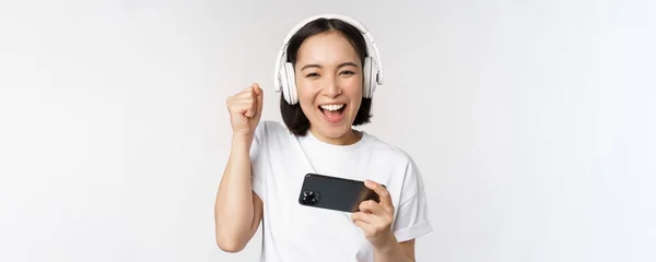Feliz chica asiática gamer, jugando en el teléfono móvil, viendo en el teléfono inteligente, usando auriculares, de pie sobre fondo blanco —  Fotos de Stock