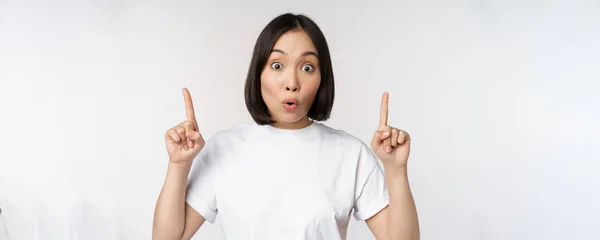 Retrato de mujer joven entusiasta, chica asiática sonriendo señalando con los dedos hacia arriba, mostrando publicidad hacia arriba, de pie sobre fondo blanco — Foto de Stock