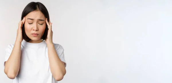 Портрет азиатской девушки с головной болью, мигренью или болезнью, стоящей в белой футболке на белом фоне — стоковое фото