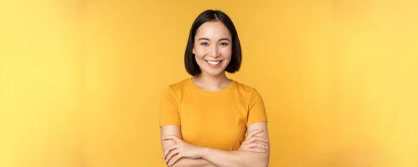 Zelfverzekerd aziatisch meisje kruis armen op de borst, glimlachend en assertief, staande over gele achtergrond — Stockfoto