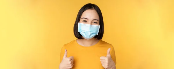 Cheerful korean woman in medical face mask, support people during pandemic, wear personal protective equipment from covid-19, showing thumbs up in approval, yellow background — Stock Photo, Image