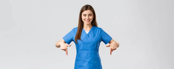 Zdravotničtí pracovníci, prevence viru, covid-19 screening, medicínský koncept. Usmívající se sebevědomá zdravotní sestra, lékař nebo stážista v modrých pláštích, ukazující prsty dolů, aby pacienti informace — Stock fotografie