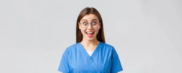 Sjukvårdspersonal, medicin, försäkring och covid-19 pandemi koncept. Entusiastiska optimistiska kvinnliga sjuksköterska, läkare i blå skurar och glasögon hör fantastiska nyheter, leende roade — Stockfoto