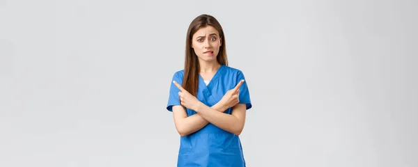 Healthcare workers, prevent virus, insurance and medicine concept. Indecisive and puzzled female nurse, doctor in blue scrubs pointing sideways at banners, biting lips nervous, need advice — Stock Photo, Image