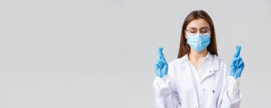 Covid-19, virüsü önleme, sağlık çalışanları ve karantina konsepti. Beyaz önlüklü umut dolu doktor, tıbbi maske ve eldivenler, kapalı gözler, çapraz parmaklar ve dua eden hastalar enfeksiyonu tedavi eder.