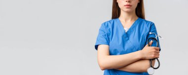 Sağlık çalışanları, ilaç, sigorta ve covid-19 salgını konsepti. Ciddi yüzlü, çapraz kollu ve steteskoplu mavi önlüklü genç bayan doktor ya da hemşire.