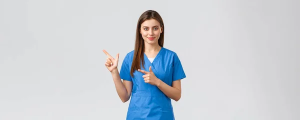 Healthcare workers, prevent virus, insurance and medicine concept. Smiling attractive female doctor or nurse in blue scrubs pointing fingers left to show patients advertisement, important info — Stock Photo, Image