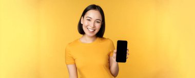 Güzel mutlu Asyalı kız cep telefonu ekranını gösteriyor, akıllı telefon cihazına başvuruyor, sarı arka planda duruyor.