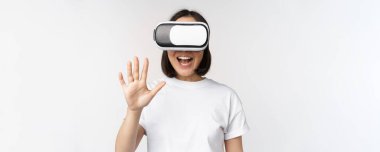 Mutlu Asyalı kadın VR kulaklık kullanıyor, el sallıyor ve gülüyor, sanal gerçeklik gözlüğü kullanıyor, beyaz arka planda duruyor.