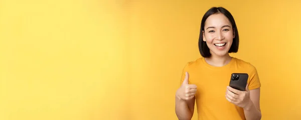 Feliz sonrisa chica asiática sosteniendo el teléfono móvil y mostrando los pulgares hacia arriba, recomendando la aplicación en el teléfono inteligente, de pie sobre el fondo amarillo —  Fotos de Stock