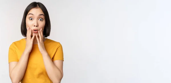 Close up portrait of asian woman looking surprised, wow face, staring impressed at camera, standing over white background in yellow t-shirt — Stock Photo, Image