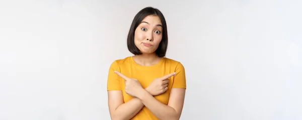 Kompliziertes asiatisches Mädchen zeigt mit dem Finger zur Seite, zeigt linke und rechte Entscheidungen, starrt unentschlossen ahnungslos über weißem Hintergrund — Stockfoto