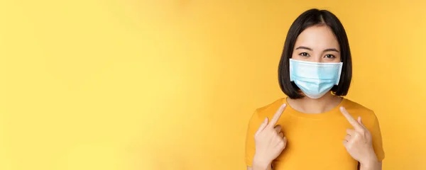 Portret van een lachende Aziatische vrouw met een medisch gezichtsmasker, wijzend naar haar persoonlijke beschermingsmiddelen van covid-19 tijdens een pandemie, staande tegen een gele achtergrond — Stockfoto