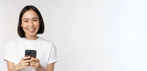 Портрет усміхненої азіатки, що використовує мобільний телефон, балачки, смс-повідомлення, стоїть у сорочці на білому тлі — стокове фото