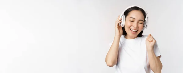 Moderna chica asiática bailando, escuchando música con auriculares, sonriendo feliz, de pie en camiseta sobre fondo blanco — Foto de Stock