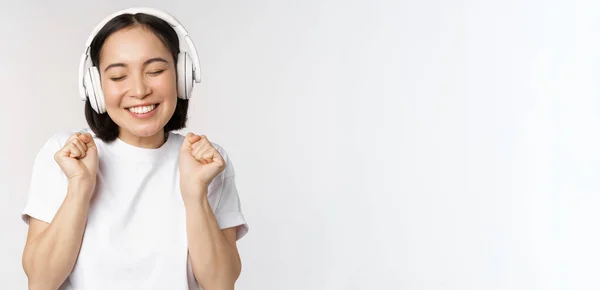 Moderna chica asiática bailando, escuchando música con auriculares, sonriendo feliz, de pie en camiseta sobre fondo blanco — Foto de Stock