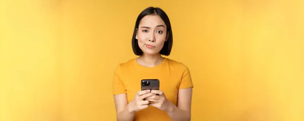 スマートフォンを持っている懐疑的なアジアの女性,カメラで疑いを持って見て,黄色の背景の上に立って — ストック写真