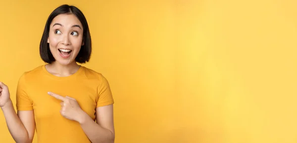 Retrato de chica asiática feliz señalando los dedos y mirando a la izquierda, sonriendo asombrado, mirando banner promocional, mostrando publicidad sobre fondo amarillo — Foto de Stock