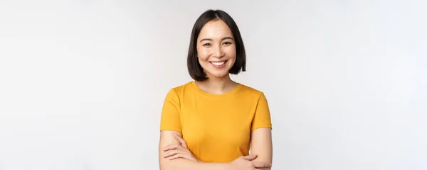 Retrato de jovem bela morena, menina asiática sorrindo e olhando feliz, cruz braços no peito, em pé sobre fundo branco — Fotografia de Stock