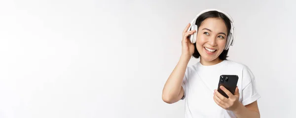 Belle fille asiatique moderne, écouter de la musique dans les écouteurs, tenant le téléphone mobile, en utilisant l'application smartphone, debout sur fond blanc — Photo