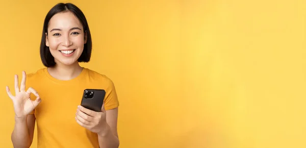Щаслива усміхнена азіатська дівчина тримає мобільний телефон і показує добре, рекомендує додаток на смартфоні, стоячи над жовтим тлом — стокове фото