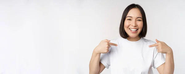 Mujer asiática feliz y confiada, estudiante sonriendo y señalándose a sí misma, autopromocionándose, mostrando el logotipo en la camiseta, de pie sobre fondo blanco — Foto de Stock
