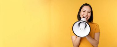 Gülümseyen Asyalı kadın megafonla duruyor, smth 'i anons ediyor, reklam ürünü, sarı arka planda duruyor.