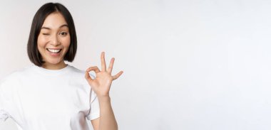 Herşey yolunda mı? Gülümseyen genç Asyalı kadın, güvence veriyor, tamam işaretini tatmin olmuş bir şekilde gösteriyor, beyaz arka planda duruyor.