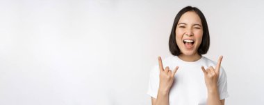 Şımarık Asyalı kız bağırıyor, konser veya festivalden zevk alıyor, rock 'ı gösteriyor, heavy metal işareti yapıyor, eğleniyor, beyaz arka planda duruyor.