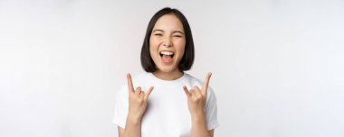 Şımarık Asyalı kız bağırıyor, konser veya festivalden zevk alıyor, rock 'ı gösteriyor, heavy metal işareti yapıyor, eğleniyor, beyaz arka planda duruyor.