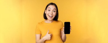 Heyecanlı Asyalı kadın cep telefonu ekranı gösteriyor, başparmaklar yukarı, tıpkı smth good gibi, akıllı telefon uygulaması öneriyor, sarı arka planda duruyor.